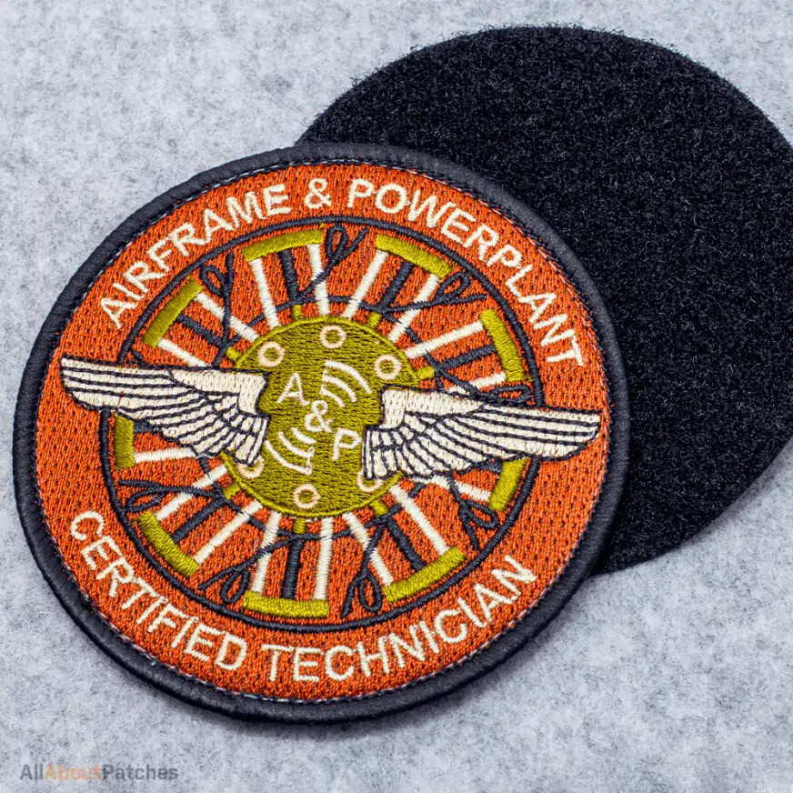 Round orange embroidered patch with winged propeller blades, beside its black hook backing on gray felt