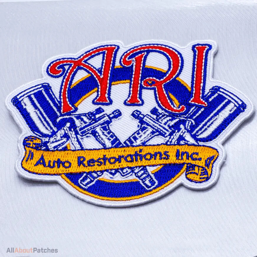 White embroidered patch with red lettering, a gold banner and crossed blue spray guns, on a silver surface