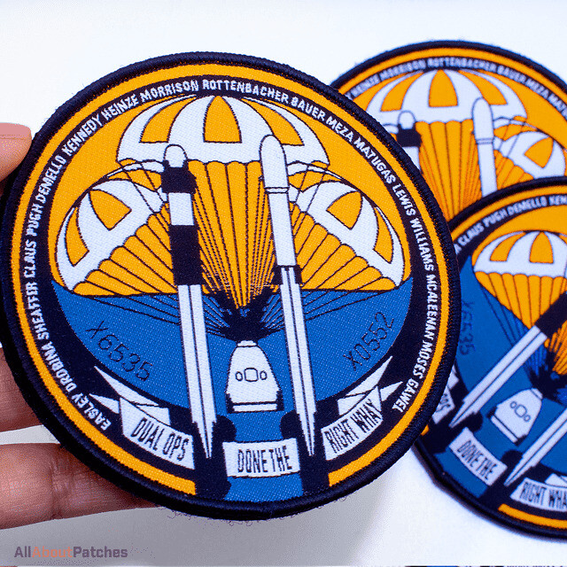 Round woven patch with rockets under orange parachutes over a blue sea, held in a hand beside two more